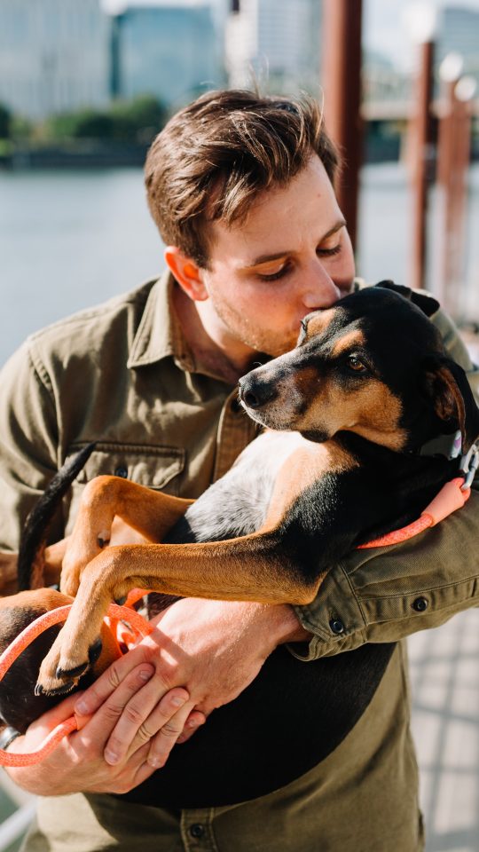 man in brown jacket hugging black and brown short coated dog